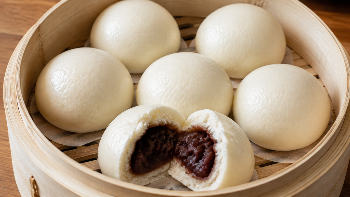 Smooth Chinese steamed buns in a bamboo steamer with one opened to show dark red bean paste